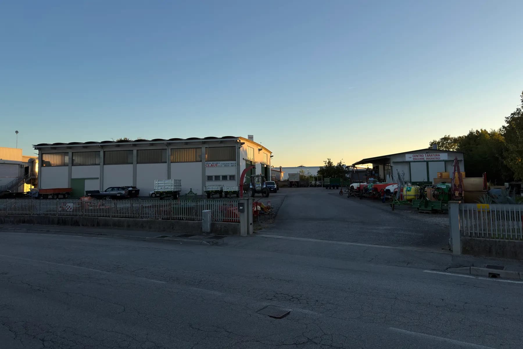 L'officina Claut a sinistra, con la scritta CLAUT sulla parete e dei rimorchi parcheggiati davanti, e a destra il capannone del centro taratura macchine irroratrici con alcune irroratrici esposte, visti dalla strada
