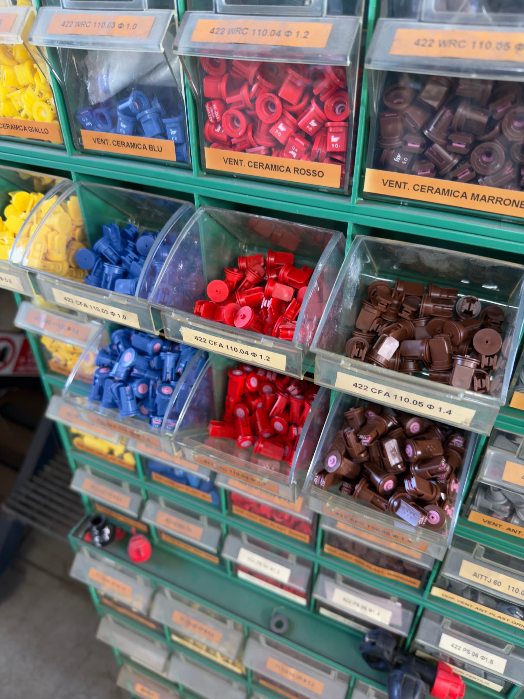 Primo piano di cassetti aperti con ugelli gialli, blu, rossi e marroni, divisi per modello e diametro con etichette come 422 CFA 110.04 e VENT. CERAMICA ROSSO