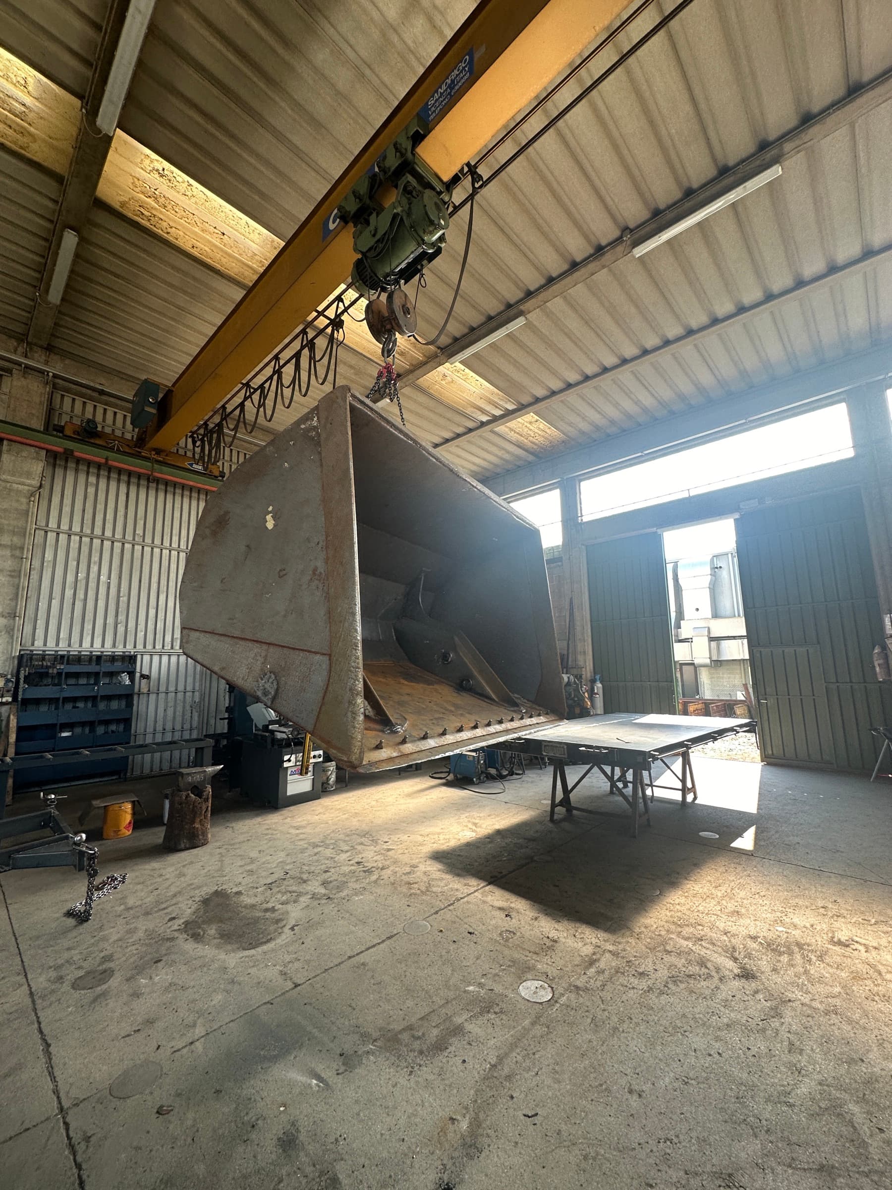 Capannone dell'officina in controluce: benna sospesa al carroponte, tavolo da lavoro su cavalletti e portone aperto sul piazzale