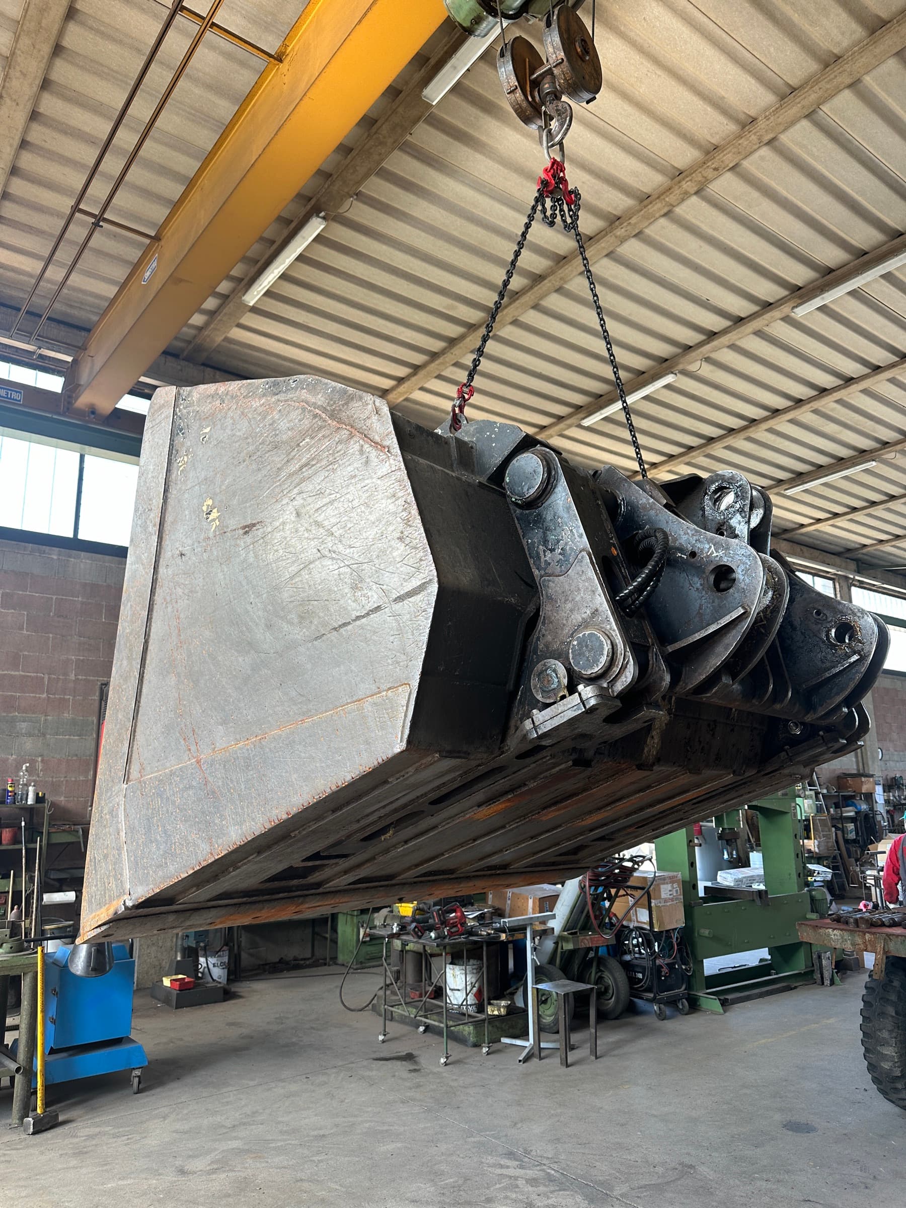 Benna da pala gommata sollevata con le catene del carroponte, vista dal basso dal lato degli attacchi, nell'officina Claut
