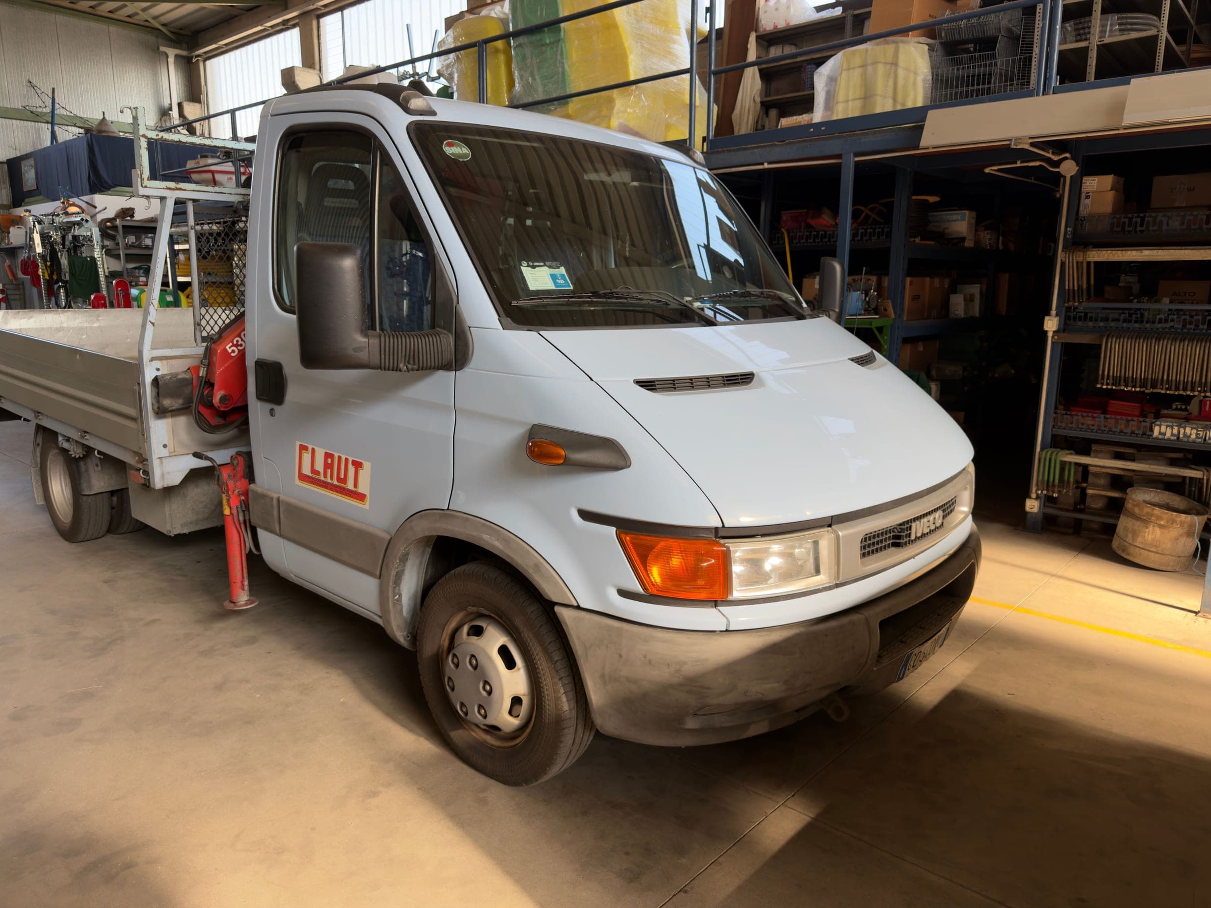 Autocarro Iveco Daily bianco con cassone e gru, col logo CLAUT sulla portiera, fermo nel capannone davanti agli scaffali